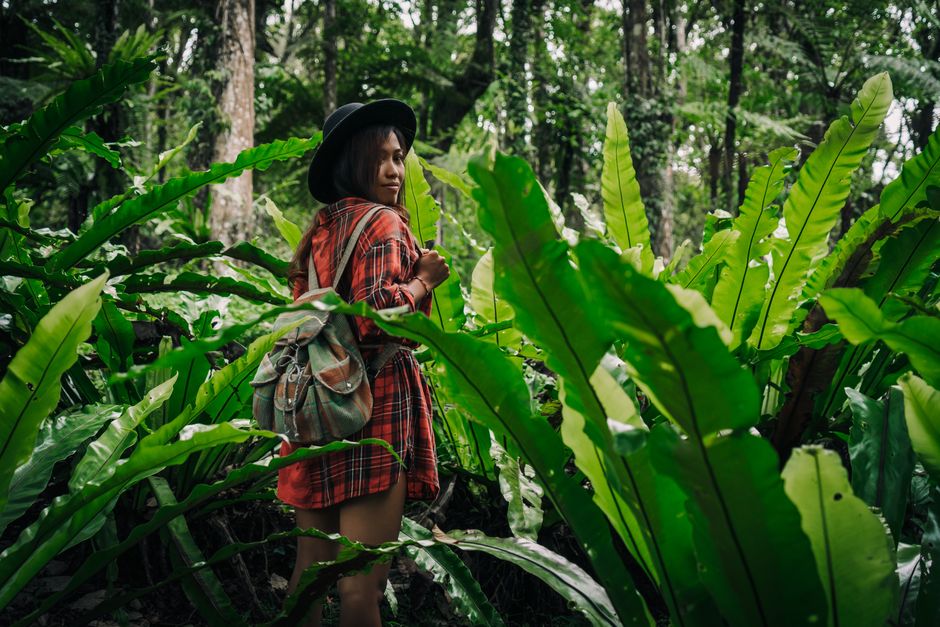 A young woman in a jungle with a backpack and hat embraces nature on a hiking adventure.