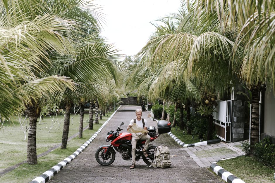 Full length male traveler in casual outfit leaning on motorcycle near duffle bag while studying city map on pavement of street among tropical trees in front of open gates of building