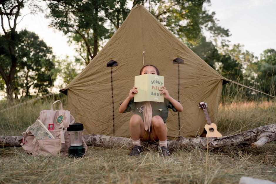 Child reading a scout field book at a campsite with a tent, ready for a camping adventure in the forest.