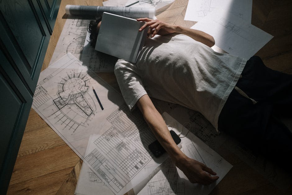 A man in casual attire resting on floor with architectural drawings and notebook.