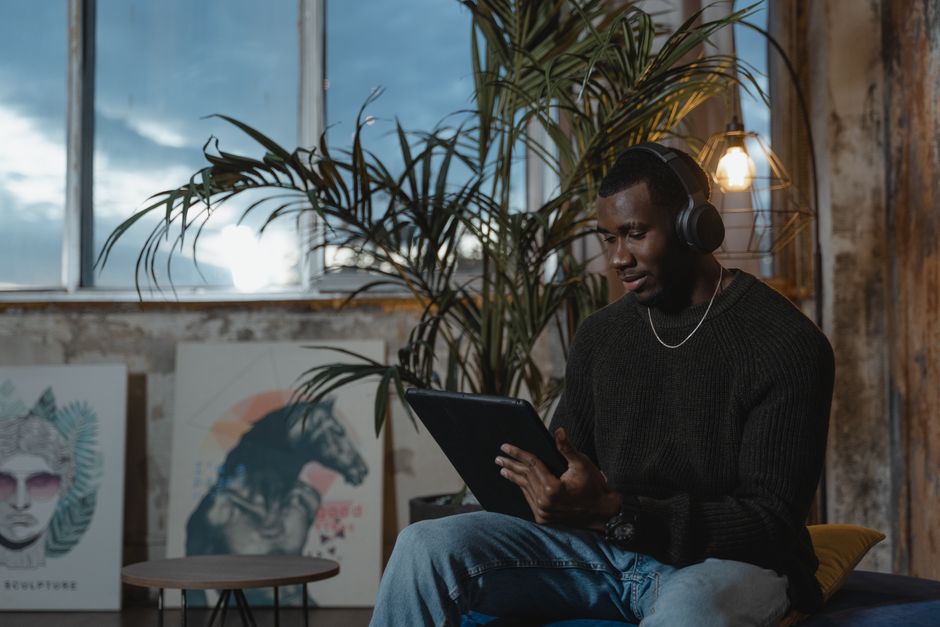 A man in a cozy room wearing headphones and using a tablet, surrounded by artwork and plants.