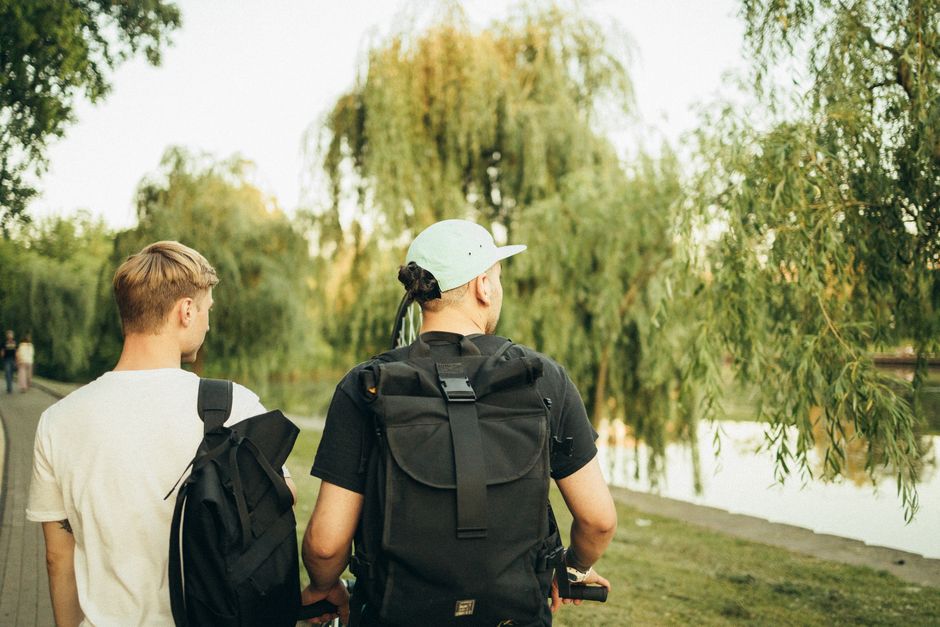 Two young men with backpacks walking in a sunny park with willow trees.