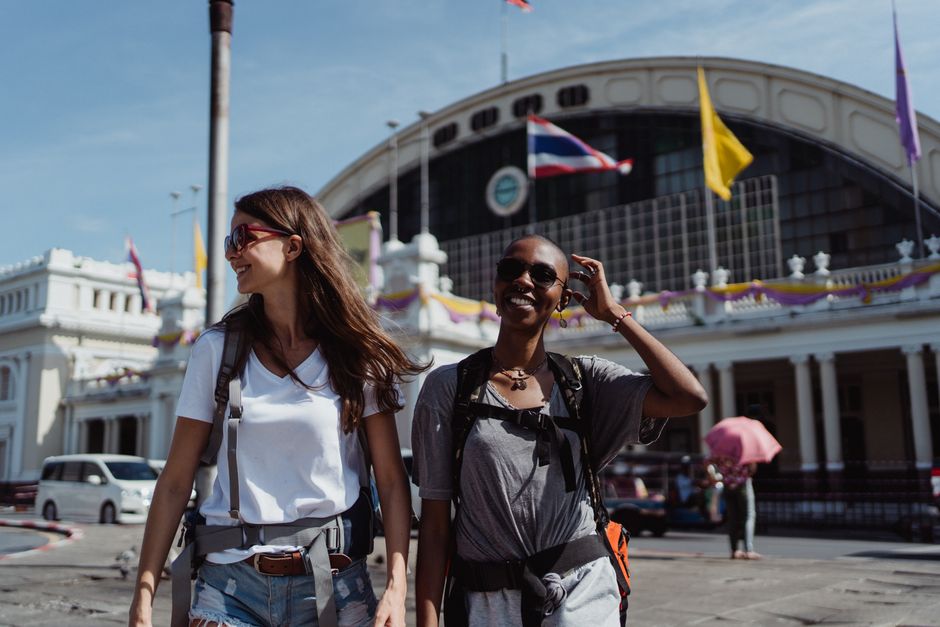 Two women walking and smiling outside in a city setting with a prominent train station in the background.