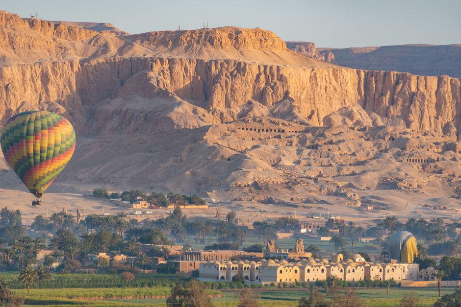 A vibrant hot air balloon glides over a beautiful Egyptian village against rugged cliffs.